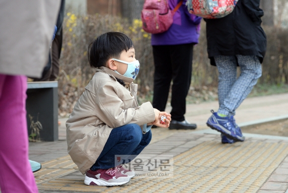 이현이가 집 앞 버스 정류장에서 외할머니와 놀이학교 버스를 기다리며 행인들 사이에 쪼그려 앉아 있다.