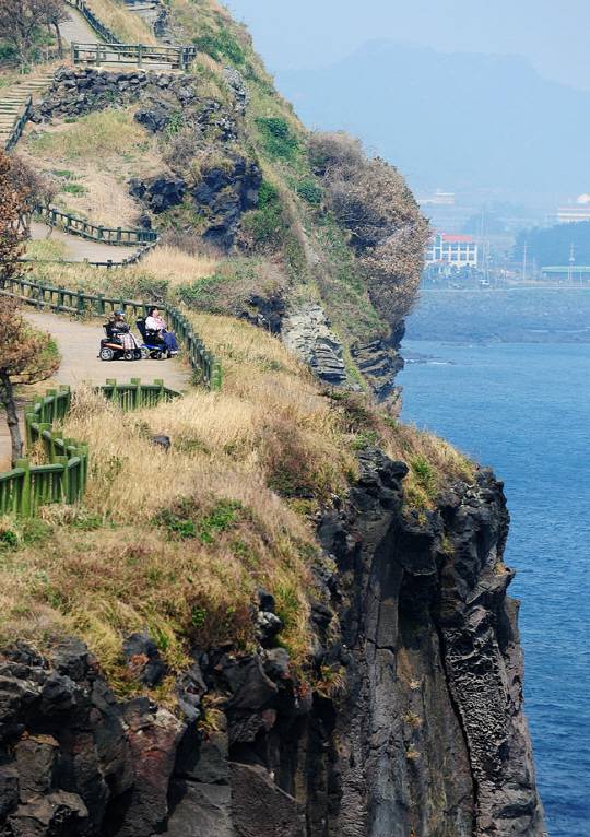 제주 송악산의 해안산책길에서 두 명의 휠체어 여행자가 벼랑 아래 쪽빛 제주 바다를 내려다보고 있는 모습.