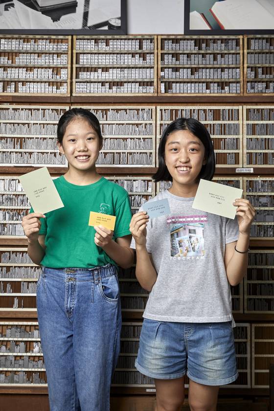 나만의 문구로 만든 활판 인쇄 엽서를 들고 활자가 가득한 문선대 앞에 선 박시은(왼쪽)·송윤서 학생기자.