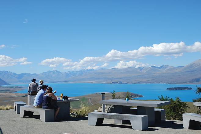 마운트 존 정상에 있는 아스트로 카페테라스에서 방문객들이 테카포호수와 파란 하늘에 늘어선 마운트 쿡 설산의 봉우리들을 조망하며 즐거운 시간을 보내고 있다.
