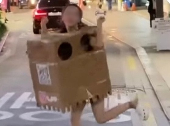 A woman in her 20s, identified as an adult performer and social media influencer who goes by the name Ain, wears a cardboard box and poses on the street of Apgujeong Rodeo in southern Seoul on Oct. 13. [SCREEN CAPTURE]