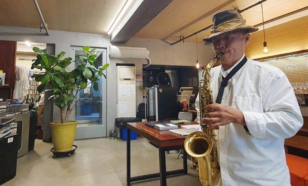 강희영 오너셰프가 그의 음식점 ‘벨 프홈나드’에서 휴식을 취하는 중 색소폰 연주를 즐기고 있다. 사진 오른쪽 중상단에 그의 애장품 하츠필드 스피커가 보인다. 강 셰프의 뒤편에는 음식점 조리대가 있다.