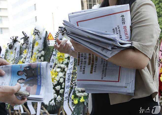 3일 오전 서울 용산구 하이브 사옥 앞에서 방탄소년단(BTS) 한 팬이 성명서가 담긴 신문을 나눠주고 있다. 그 뒤로는 BTS 지지모임이 보낸 근조화환이 세워져 있다. 이들은 하이브에 소속 아티스트 BTS 명예훼손 법적절차 진행을 촉구했다.  ⓒ News1 권현진 기자