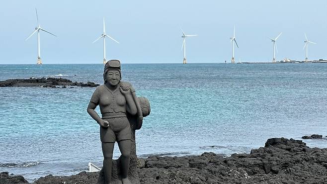 지난 3월29일 제주도 한경면 해안의 해녀 석상 뒤로 탐라해상풍력발전단지의 풍력발전기들이 돌아가고 있다. 제주도는 풍력과 같은 재생에너지 발전을 늘려 국가 탄소중립 목표 연도인 2050년보다 15년 이른 2035년에 탄소중립에 도달하겠다는 계획이다. 연합뉴스