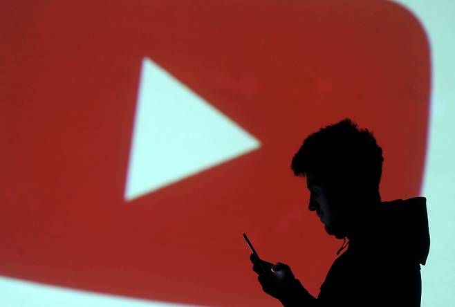 유튜브 로고를 배경으로 한 남성이 스마트폰을 하는 모습. /로이터 연합뉴스