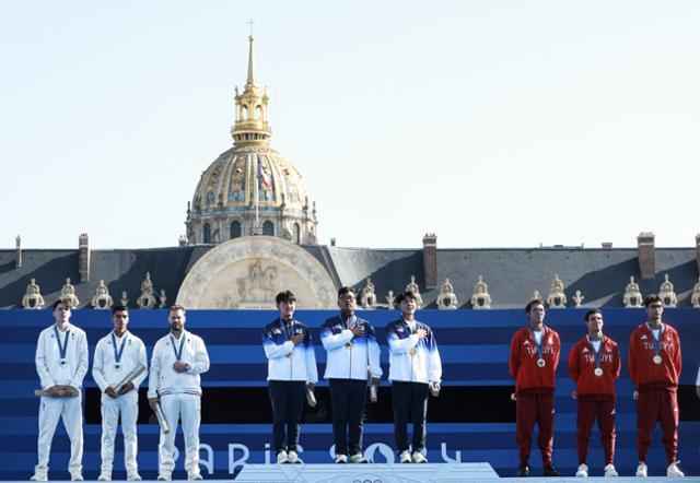 대한민국 양궁 대표팀 김우진, 김제덕, 이우석이 30일(한국시간) 프랑스 파리 앵발리드에서 열린 2024 파리 올림픽 양궁 남자 단체전 결승 시상식에서 애국가가 울려퍼지자 가슴에 손을 올리고 있다. 한국 선수들 뒤쪽으로 앵발리드의 황금 돔이, 왼쪽으로 은메달을 딴 프랑스 대표팀이 서 있다. 파리=뉴스1