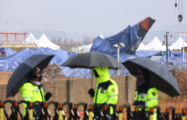 5일 전남 무안국제공항 제주항공 여객기 참사 현장에서 빗속에 우산을 쓰고 근무하는 경찰관들 뒤로 사고기 꼬리 날개에 방수포가 덮여 있다. 연합뉴스