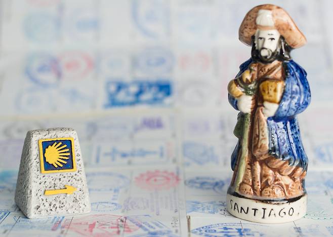 Symbols of the Way of Saint James. Miniature of a sign mark with yellow arrow and Statuette of St James (Santiago apostle). Pilgrim‘s passport filled with stamps on background