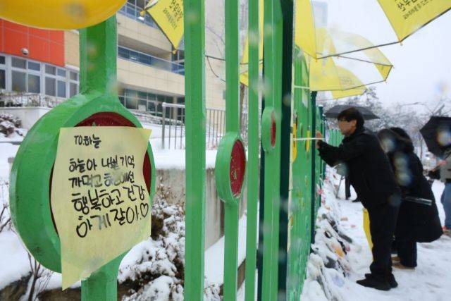 대전 초등학생 김하늘양 피살사건이 발생한 대전 서구의 한 초등학교 정문에서 12일 오전 학교 관계자가 추모객들이 놓고 간 꽃과 편지 위에 우산을 씌워주고 있다.ⓒ연합뉴스