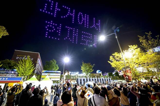 군산 시간여행축제에서 드론이 밤하늘에 슬로건을 수놓자 관광객들이 일제히 휴대전화 카메라에 담고 있다. 군산시 제공