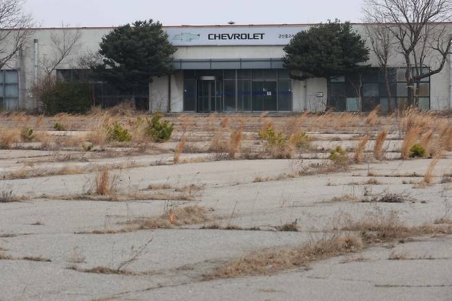 16일 전북 군산시 한국GM 군산공장 출고 대기장의 모습. 굳게 닫힌 녹슨 철문 뒤로 잡초가 무성한 빈 땅과 멀리 빛바랜 쉐보레 로고가 보인다. [한주형 기자]