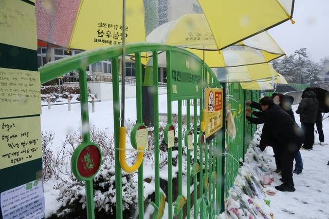 ▲ 대전 초등학생 김하늘 양 피살사건이 발생한 대전 서구의 한 초등학교 정문에서 지난 2월 12일 오전 학교 관계자가 추모객들이 놓고 간 꽃과 편지 위에 우산을 씌워주고 있다. 학교 정문에는 시민들이 붙여놓은 쪽지와 꽃, 인형, 선물들이 가득 차 있는 모습이다.  연합뉴스