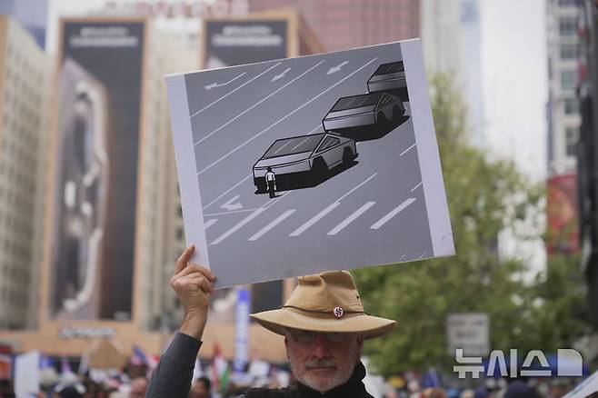 [로스앤젤레스=AP/뉴시스] 1일(현지 시간) 미 캘리포니아주 로스앤젤레스에서 열린 노동절 시위에 참여한 한 남성이 테슬라 자동차에 반대하는 내용의 그림을 들고 있다. 2025.05.02.