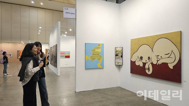 ‘아트부산 2025’ 전경. 두 관람객이 갤러리조은 부스에 걸린 일본작가 마이코 코바야시의 ‘둘’(2022)을 바라보며 환하게 웃고 있다. 옆으로 회화작품을 설치작품처럼 내걸어 시선을 끈 이재현의 ‘당신의 동화가 여기 있습니다’(2025)와 백윤조의 ‘무임승차자’(2025)가 보인다(사진=오현주 문화전문기자).