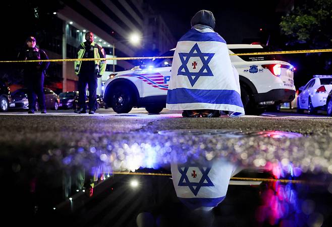 21일 미국 워싱턴 DC의 수도 유대인 박물관 근처에서 이스라엘 대사관 직원 2명이 총격을 받아 사망한 현장에서 이스라엘 국기를 두른 한 남성이 폴리스라인 밖에 앉아 있다./로이터 연합뉴스