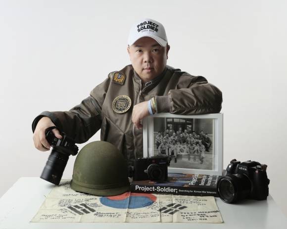 라미 현 작가가 그의 작품 사진과 작업용 카메라, 철모, 한국전 때 사용됐던 태극기와 함께 포즈를 취하고 있다. 라미 현 작가 제공