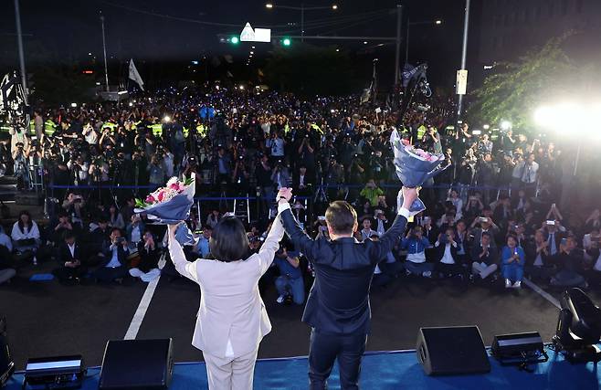 제21대 대통령 당선이 확실시되는 더불어민주당 이재명 대선 후보와 부인 김혜경 여사가 4일 서울 여의도 국회 인근에서 당 주최로 열린 국민개표방송 행사에 참석해 꽃다발을 받고서 시민들을 향해 두 팔을 들어 인사하고 있다. [연합]