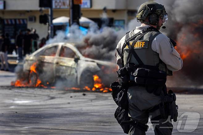 미국 이민세관단속국(ICE)이 7일(현지시간) 캘리포니아주 로스앤젤레스 카운티의 파라마운트에서 불법 체류자 체포 후 시위대와 경찰이 대치하는 가운데 도로 위 불타는 차량에서 연기가 피어오르고 있다. 2025.6.7./뉴스1 ⓒ 로이터=뉴스1 ⓒ News1 이창규 기자