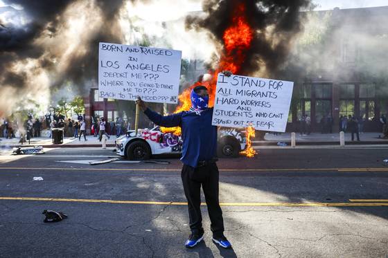 현지시간 8일 미국 캘리포니아주 로스앤젤레스에서 이민 단속으로 촉발된 시위에서 한 시위 참석자가 불타는 차량 앞에서 팻말을 들고 있다. EPA=연합뉴스