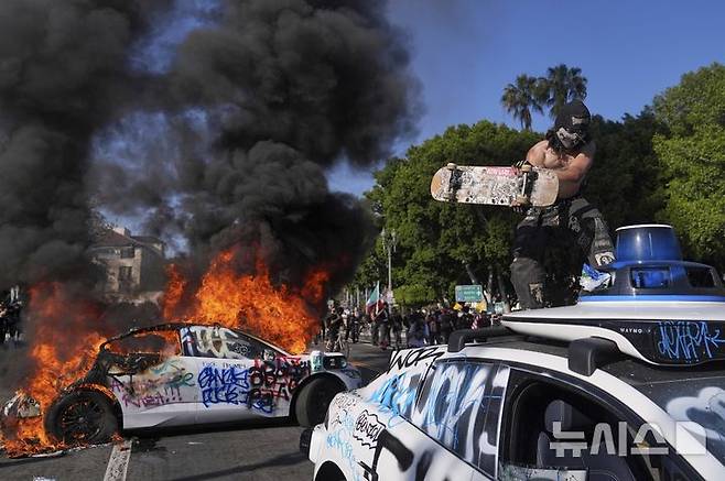 [로스앤젤레스=AP/뉴시스] 8일(현지 시간) 미 캘리포니아주 로스앤젤레스 도심 메트로폴리탄 구치소 인근에서  무인 자율주행 택시 웨이모가 불타는 가운데 한 시위 참가 남성이 또 다른 차량을 훼손하고 있다. 2025.06.09.