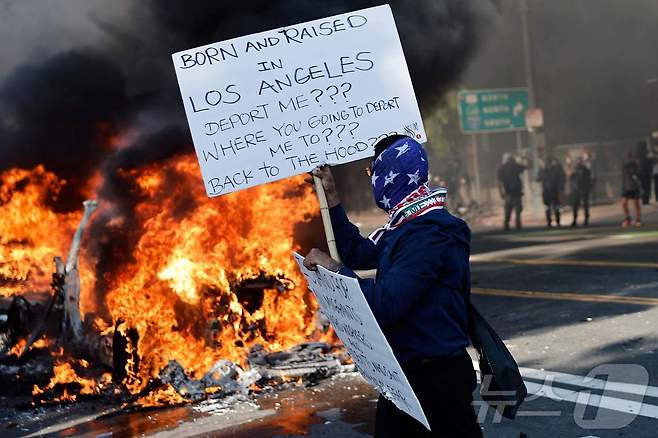 2025년 6월 8일 캘리포니아주 로스앤젤레스(LA)에서 사람들이 불타는 웨이모(Waymo) 차량 옆에 서 있다. 이민 단속 과정에서 경찰과의 충돌이 이틀간 이어진 후, 도널드 트럼프 행정부가 캘리포니아주의 반대에도 불구하고 주방위군을 투입하면서 도시 내 긴장이 고조되고 있다. 2025.6.8 ⓒ AFP=뉴스1 ⓒ News1 강민경 기자