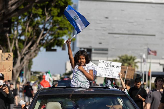 가족 지키려, 소녀도 나섰다 도널드 트럼프 미국 행정부의 이민자 단속에 항의하는 시위대가 11일(현지시간) 캘리포니아주 로스앤젤레스(LA) 도심 근처를 행진하는 가운데 한 소녀가 차량 루프로 몸을 내밀어 엘살바도르 국기를 흔들고 있다. AFP연합뉴스