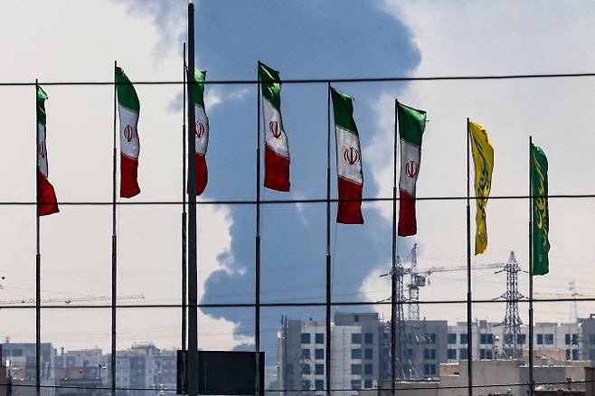 15일(현지시간) 밤사이 이스라엘의 공습으로 타격을 입은 남부 테헤란의 정유소에서 짙은 연기와 불길이 치솟는 가운데, 다리 위에는 여러 국기가 펄럭이고 있다. (사진=  AFP)