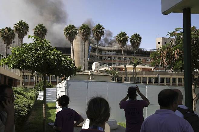 지난 19일(현지 시간) 이스라엘 베르셰바에서 시민들이 이란의 미사일 공격으로 연기가 치솟는 소로카 병원을 지켜보고 있다. 이란 혁명수비대는 “병원 근처 이스라엘군 지휘·정보센터가 타격받았다”고 주장했다. AP·뉴시스