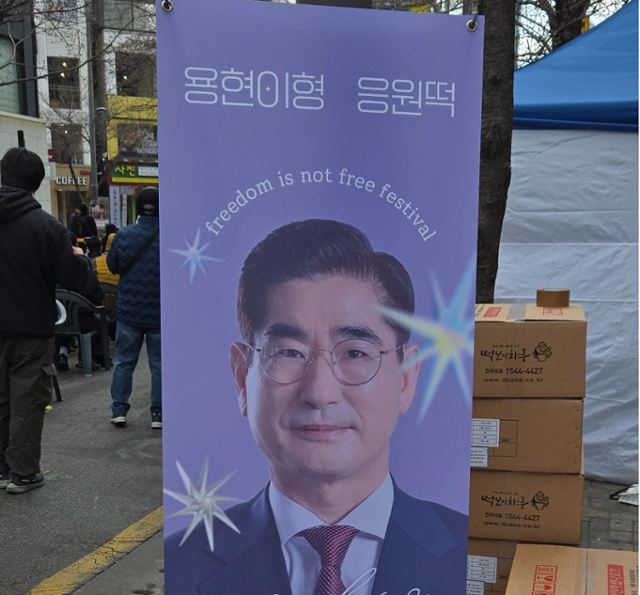 3월 29일 서울 서대문구 신촌역 일대에서 '탄핵을 반대하는 대한민국 청년' 등 주최로 열린 자유 토크쇼 행사장에 김용현 전 국방부 장관이 지지자들에게 '응원떡'을 돌린다는 내용을 적은 입간판이 설치돼 있다. 엑스(X) 캡처