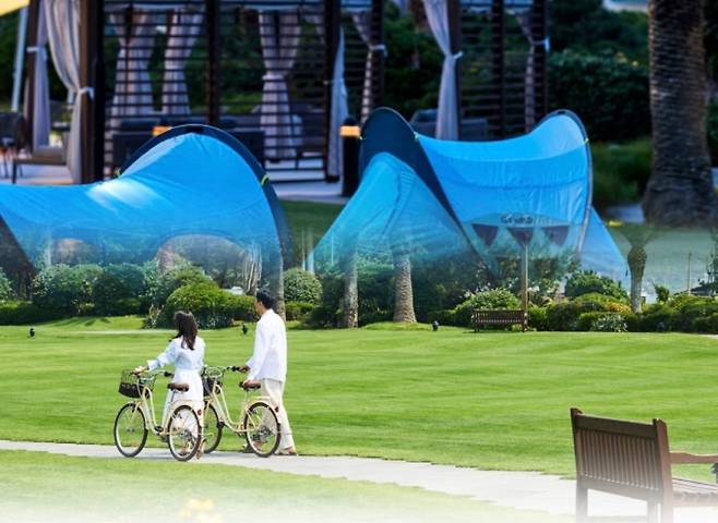 자전거를 끌고 정원을 걷는 투숙객(아래)과 바다를 마주한 잔디 위 쉼터 텐트. 제주 관광이 ‘체류 중심’으로 옮겨가는 흐름을 상징적으로 보여준다.