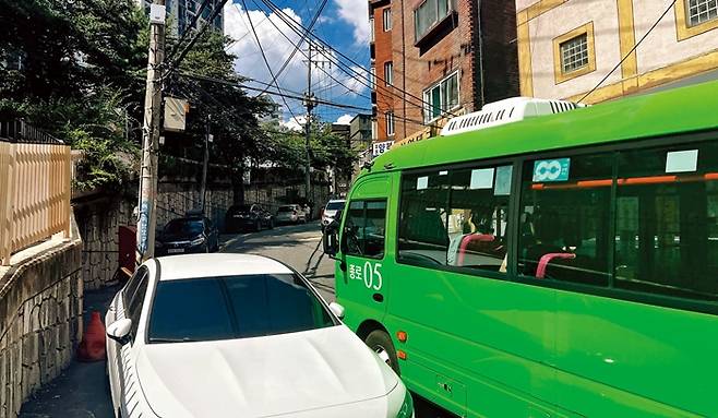 마을버스가 좁은 이면도로에 불법주차된 승용차를 아슬아슬하게 피해 언덕을 오르고 있다.