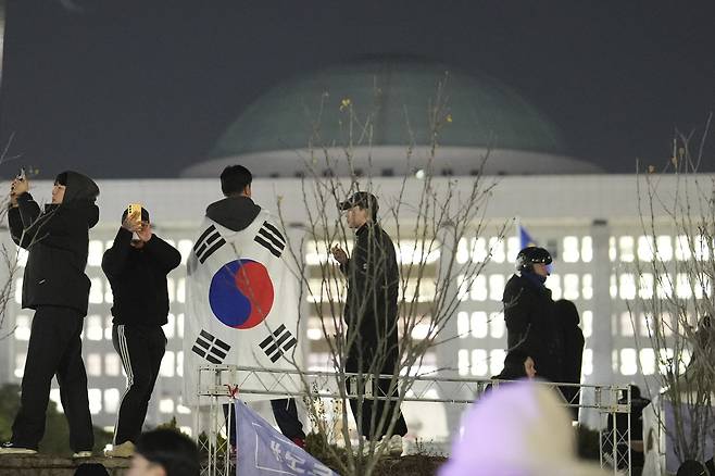 A man with the Korean flag draped over his shoulders stands on the wall of the National Assembly in Yeouido, western Seoul, in the early hours of Dec. 4, 2024. [AP/YONHAP]