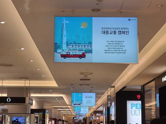 천장에 매달린 광고판에는 롯데타워와 2층 버스 그림이 있고, ‘활대차원 절감과 함께하는 대중교통 캠페인’이라는 텍스트가 쓰여 있는 사진이다.광고판 아래로 쇼핑몰 내부가 길게 뻗어 있는 모습이 보인다.