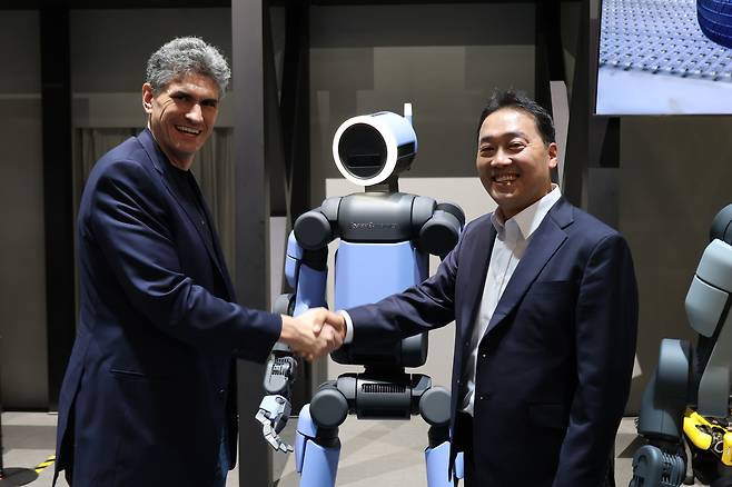 Zachary Jackowski (left), head of Atlas development at Boston Dynamics, and Oh Se-wook, executive director of steering and suspension R&D at Hyundai Mobis, shake hands in front of Boston Dynamics’ Next-Generation Electric Atlas Production Model before a group interview at the Las Vegas Convention Center on Wednesday. (Hyundai Motor Group)