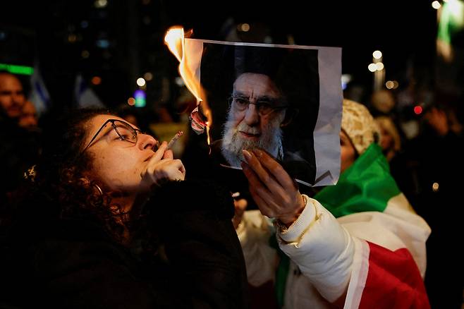 A woman lights a cigarette with fire from a burning picture of Iran's Supreme Leader Ayatollah Ali Khamenei, as Israelis rally in support of the nationwide protests happening in Iran, in Holon, Israel, on Jan. 14. [REUTERS/YONHAP]