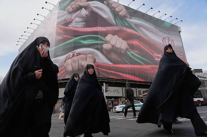 Women cross a street under a huge banner showing hands firmly holding Iranian flags as a sign of patriotism, as one of them flashes the victory sign, in Tehran, Iran, on Jan. 14. [AP/YONHAP]