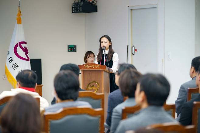 연단에서 발표를 하고 있는 여성과 청중들의 모습이 담긴 사진이다. 여성은 마이크가 달린 연단 앞에서 서서 발표를 하고 있으며, 청중들은 의자에 앉아 발표를 듣고 있다.