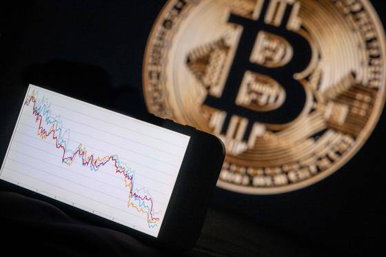 This illustration photograph shows a phone with a downward trend in the stock market in front of a screen displaying a Bitcoin logo, in Mulhouse on Dec. 5, 2025. [AFP/YONHAP]