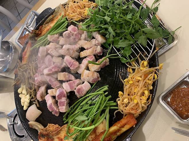 솥뚜껑 불판 위에서 두툼한 삼겹살이 노릇하게 익어가는 모습. 미나리와 콩나물, 고사리, 김치 등 다양한 채소가 곁들여져 있다. 게티이미지