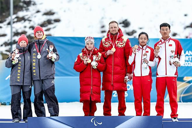 독일의 린 카즈마이어(왼쪽)가 가이드 플로리안 바우만과 함께 11일 이탈리아 테세로 크로스컨트리 스타디움에서 열린 2026 밀라노·코르티나담페초 동계 패럴림픽 여자 크로스컨트리 클래식 시각장애 스프린트 시상식에서 은메달을 목에 건 뒤 금메달을 따낸 러시아 선수들과 거리를 둔채 포즈를 취하고 있다. 타스통신