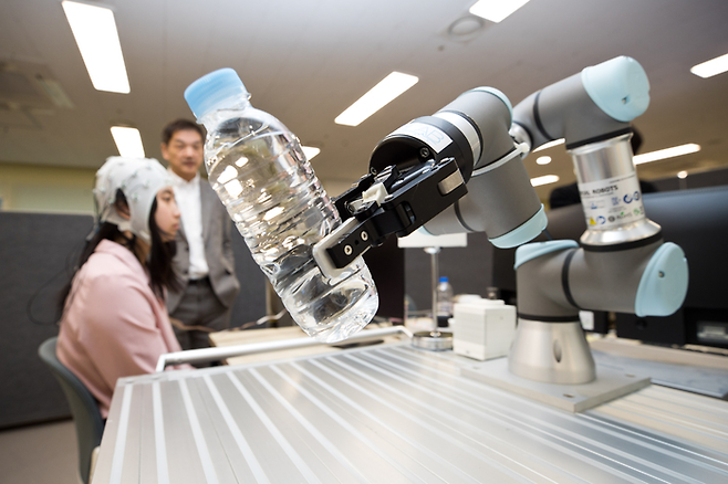 뇌-컴퓨터 인터페이스(BCI) 장비를 머리에 착용한 시연자가 물병을 잡겠다고 생각을 하자 로봇 팔이 물병을 잡아 들어올리고 있다. BCI 장비 착용자의 뇌파를 읽고, 이 신호를 로봇팔에 전달하는 데 성공한 것이다. 실제 뇌에 부착할 경우 정확도는 더 올라간다고 한다. [과학기술정보통신부]