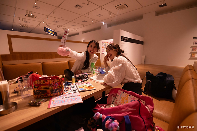 헤이세이 시절, 여학생들이 방과 후 패밀리레스토랑에서 노는 방식을 재현한 공간에서 사람들이 피처폰으로 사진을 찍고 있다. [헤이세이 연애전]