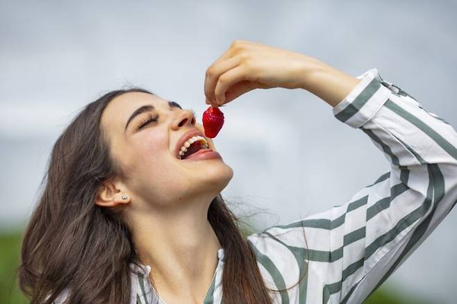 젊은 여성이 딸기를 입 안에 넣으려고 하고 있다. 딸기와 블루베리 등 플라보노이드가 풍부한 과일을 꾸준히 섭취한 사람일수록 행복감과 낙관성을 더 오래 유지하는 경향이 관찰됐다. 사진=게티이미지뱅크