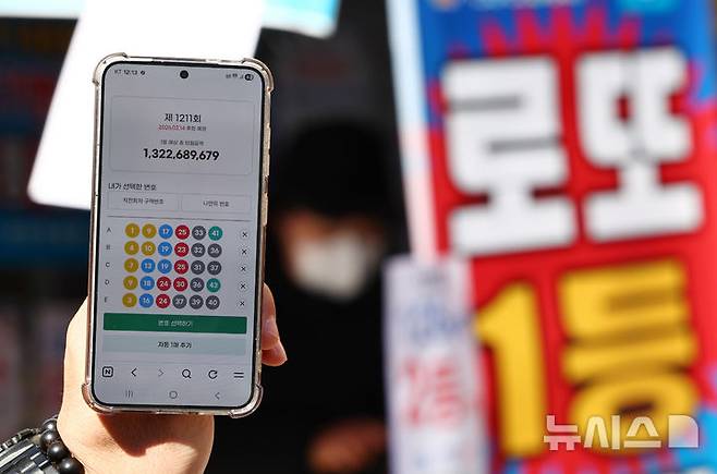 [서울=뉴시스] 김명년 기자 = 사진은 지난 2월9일 오전 서울 시내 한 복권 판매점 앞에서 휴대폰 화면에 나오는 로또 모바일 판매 페이지 뒤로 시민들이 로또를 구매하고 있는 모습. 2026.02.09. kmn@newsis.com