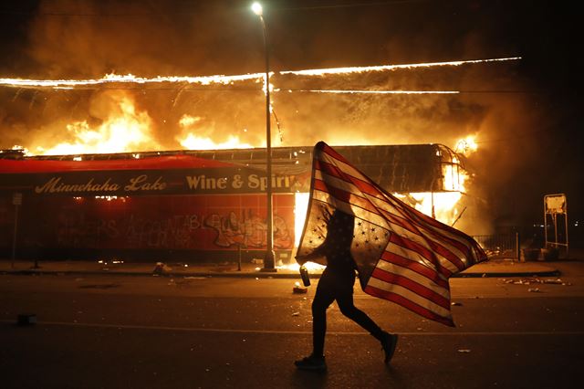 경찰의 흑인 과잉진압에 항의하는 한 시위자가 28일 미국 미네소타주 미니애폴리스에서 불에 타고 있는 건물 앞으로 미국 국기를 들고 지나가고 있다. 미니애폴리스=AP 연합뉴스