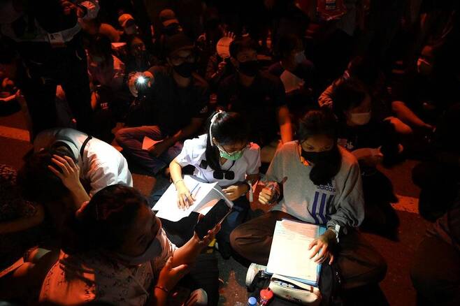 반정부 시위에 참여한 태국 여학생들이 땅바닥에 앉아 주변에서 비춰주는 불빛 도움을 받아 학교 숙제를 하는 모습.   [카오솟 캡처. 재판매 및 DB 금지]