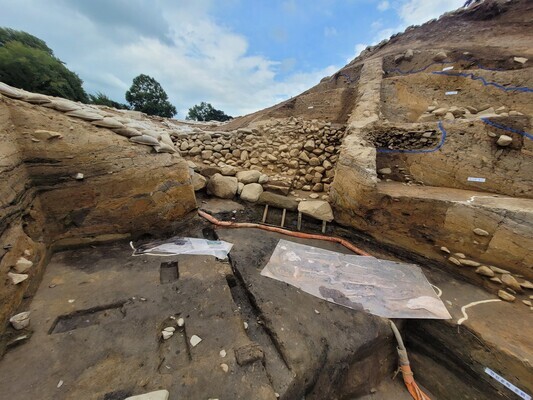 Taken on Sept. 7, this photo shows the basal soil layer blackened in the “leaf mat method” of engineering in which a layer of rice straw was burned on it, and two sheets of paper indicating where human remains were found in 2017 and this year. Above the excavation site, the palace’s central structure could be seen, with large and long stones placed in a row with a layer of additional stones over top. Next to that central structure, there were marks of filling with an additional layer of soil to increase the wall’s width. (provided by Gyeongju National Research Institute of Cultural Heritage)