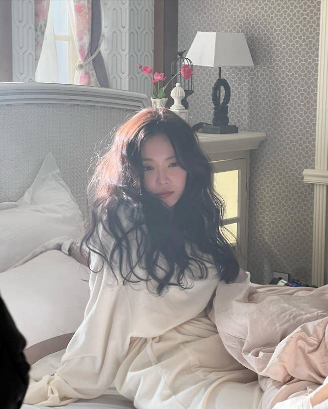 In the photo, Son Na-eun sits on the bed and looks as if she has just woken up. She has a pure charm in a loose white gown with a tangled wave hair.In another photo, she was sitting in an armchair and showed a cute and lovely face with a playful look.
