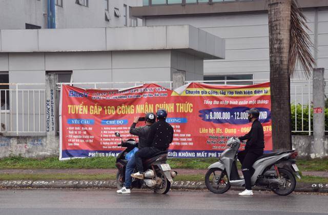 오토바이를 탄 베트남 청년들이 지난 21일 박장성 꽝쩌우 산업단지에 위치한 홍콩 디스플레이 제조기업 블루웨이 테크놀로지 베트남 생산공장 앞에 걸린 인력 모집 현수막 사진을 찍고 있다. '24시간 에어컨 가동' 등의 내용이 담겼다. 박장=허경주 특파원