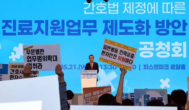 박민수 보건복지부 제2차관이 21일 서울 용산구 피크앤파크 컨벤션 로얄홀에서 열린 간호법 제정에 따른 진료지원업무 제도화 방안 공청회에 참석해 개회사를 하는 가운데 전국민주노동조합총연맹 공공운수노조 의료연대본부 조합원이 항의하고 있다./연합뉴스